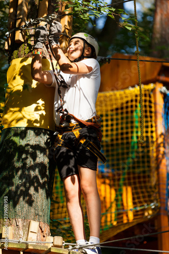 Child in forest adventure park. Kid in white helmet and white t shirt climbs on high rope trail. Agility skills and climbing outdoor amusement center for children. young boy plays outdoors