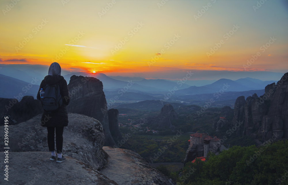 Fototapeta premium Happy woman stay on the peak of the mountain cliff edge under sunset light sky enjoying the success, freedom and bright future. Monastery Meteora Kalambaka in Greece on background.