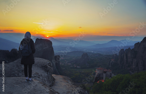 Happy woman stay on the peak of the mountain cliff edge under sunset light sky enjoying the success, freedom and bright future. Monastery Meteora Kalambaka in Greece on background.