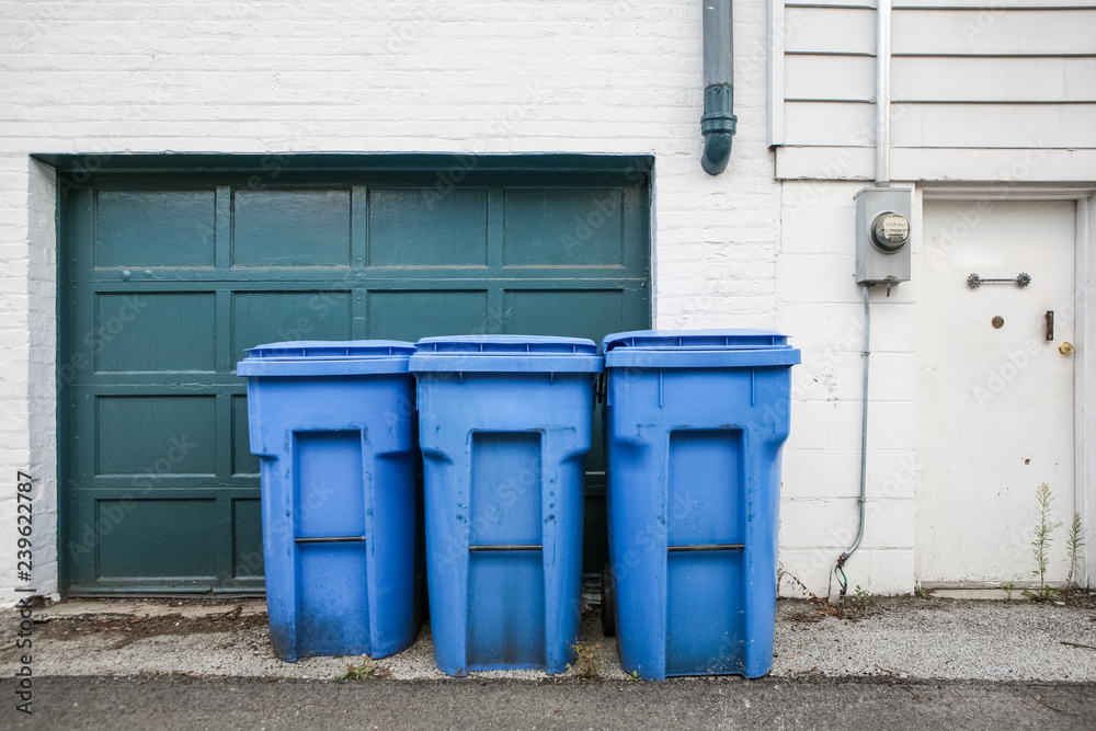 Three blue trash cans with wheels by a garage door in an alley Three blue trash cans with wheels by a garage door in an alley