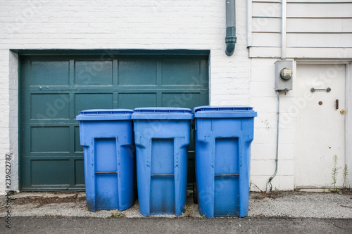 Three blue trash cans with wheels by a garage door in an alley