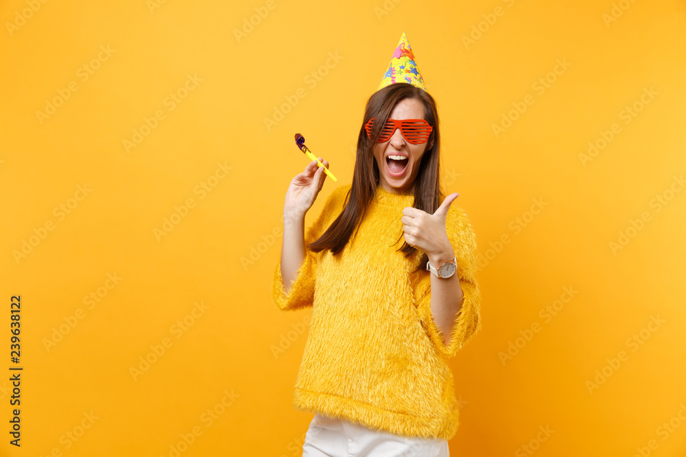 Cheerful woman in orange funny glasses, birthday hat with playing pipe showing thumb up, celebrating isolated on bright yellow background. People sincere emotions lifestyle concept. Advertising area.