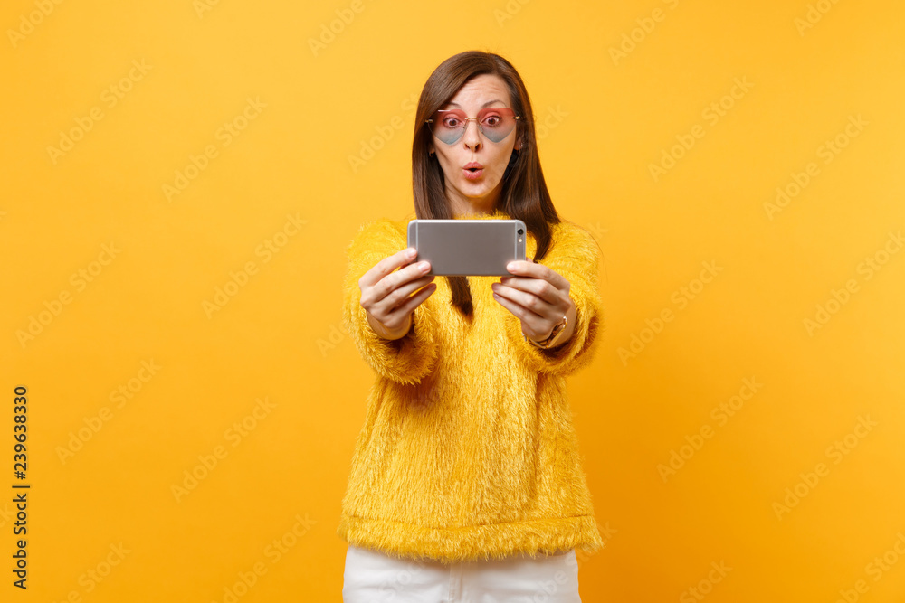 Amazed pretty young woman in fur sweater and heart glasses doing taking selfie shot on mobile phone isolated on bright yellow background. People sincere emotions, lifestyle concept. Advertising area. Amazed pretty young woman in fur sweater and heart glasses doing taking selfie shot on mobile phone isolated on bright yellow background. People sincere emotions, lifestyle concept. Advertising area.