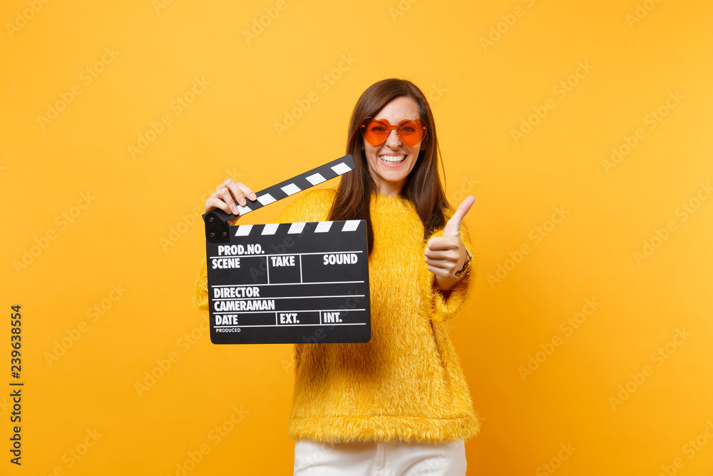 Smiling young woman in orange heart eyeglasses showing thumb up, holding classic black film making clapperboard isolated on yellow background. People sincere emotions, lifestyle. Advertising area.