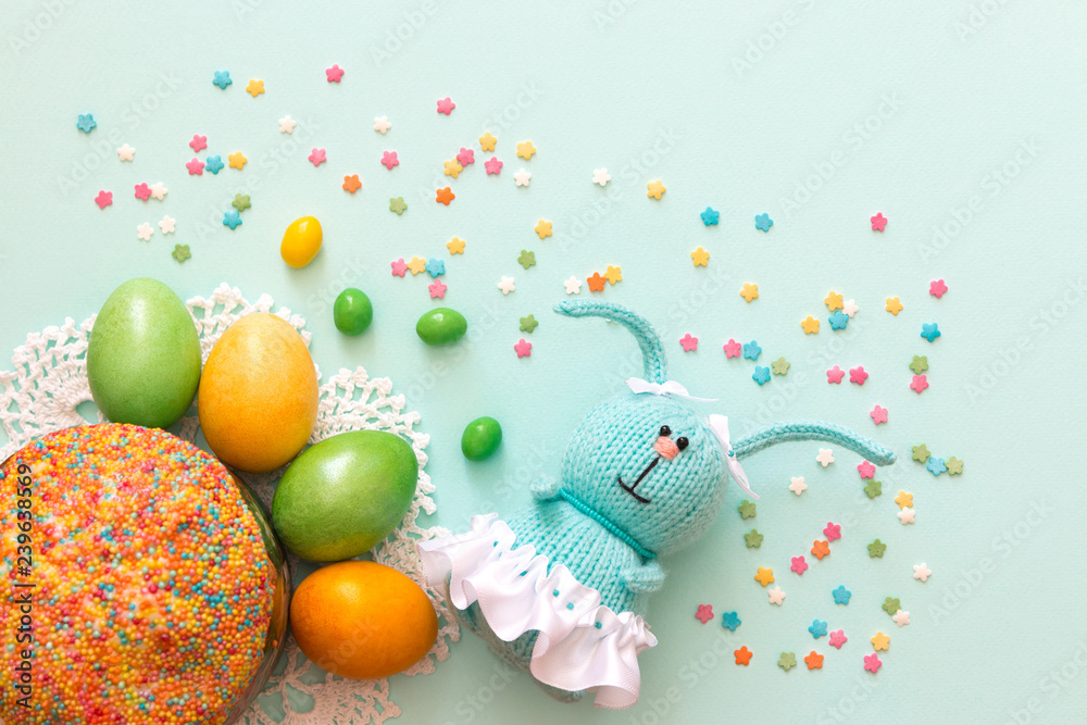 Easter celebration. Cute handmade knitted hare - a symbol of the holiday. Near her are colored eggs and Easter cake. Free space for text. Greeting card.