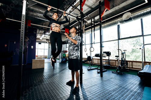 Young athletic man helps slender pretty girl to do pull up on the bar in the gym