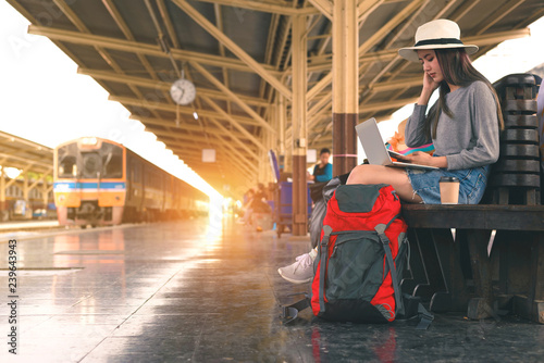 Young woman  freelance working laptop and using mobile for searching location at train station before travel, Summer travel,hipster travel are Waiting for the train at the platform of railway station.