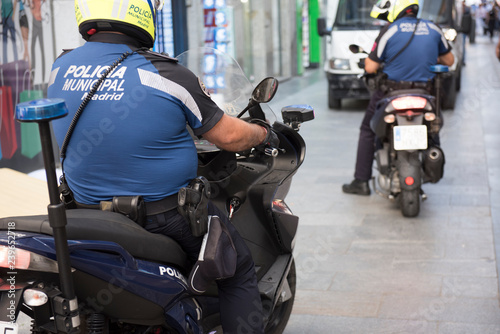 In this photo, a overweight policeman wearing his uniform is seen riding a bike. He is seen wearing a helmet and a stick attached to his back. On the background, other traffic are seen.