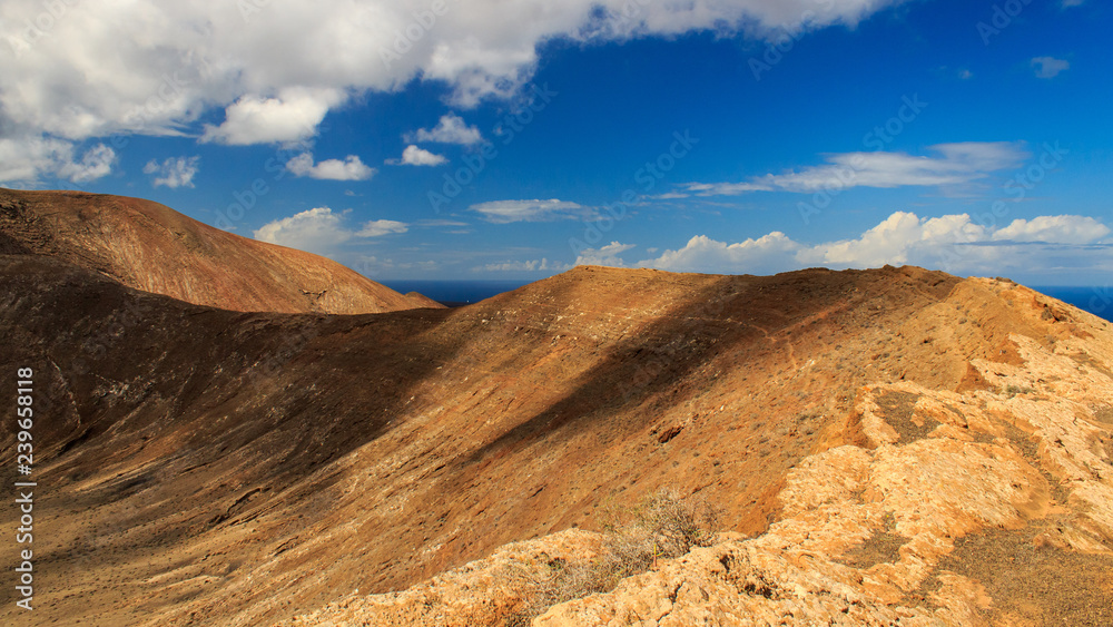 Caldera Blanca volcano crater edge on a sunny summer day under a vibrant blue sky with white clouds. Arid volcanic landscape in Lanzarote, Canary Islands, Spain.