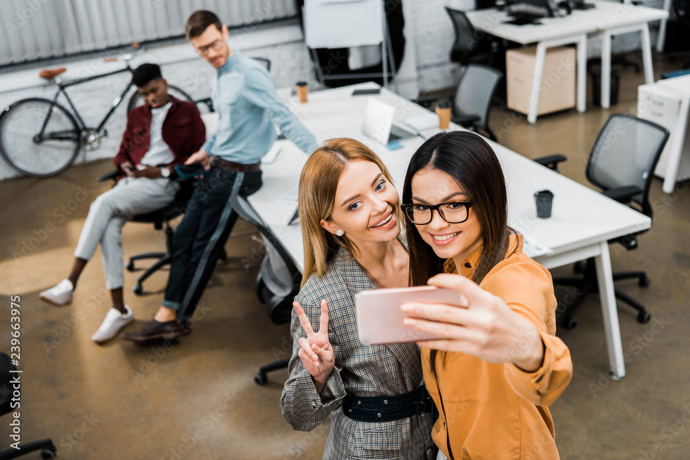 Fototapeta premium smiling businesswomen taking selfie on smartphone in office with multiracial colleagues behind