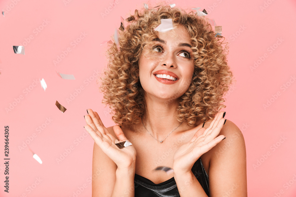 Fototapeta premium Photo of european curly woman 20s wearing dress smiling while standing under falling confetti, isolated over pink background