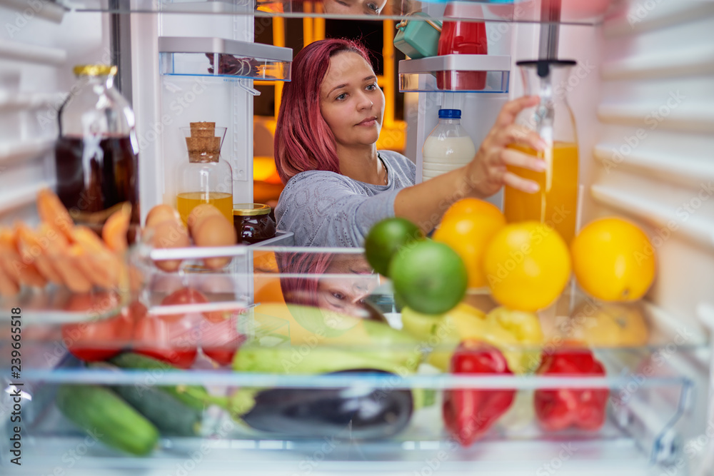Thick Caucasian woman with red hair taking bottle with juice from fridge full of groceries. Picture taken from the inside of fridge.