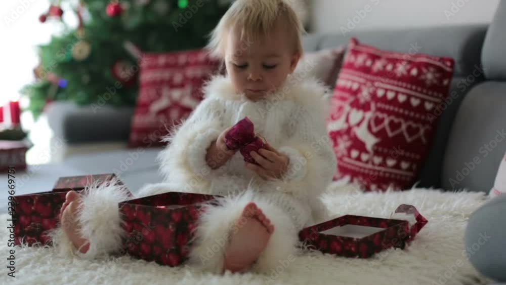 Curious little toddler boy, dressed in handknitted white overall, lying on the couch playing with gifts and happily smiling Curious little toddler boy, dressed in handknitted white overall, lying on the couch playing with gifts and happily smiling