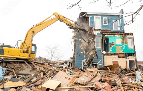 Demolishing a building with a large backhoe. Overcast day. Air is dusty. Backhoe is on debris from the demolition. Debris falls from a wall being demolished.