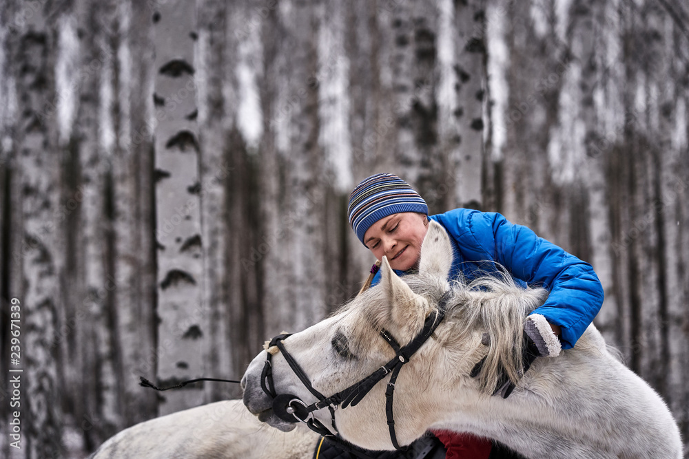 Fototapeta premium Young pretty woman in a blue jacket and a sports hat for a walk with a white horse in the winter birch forest.