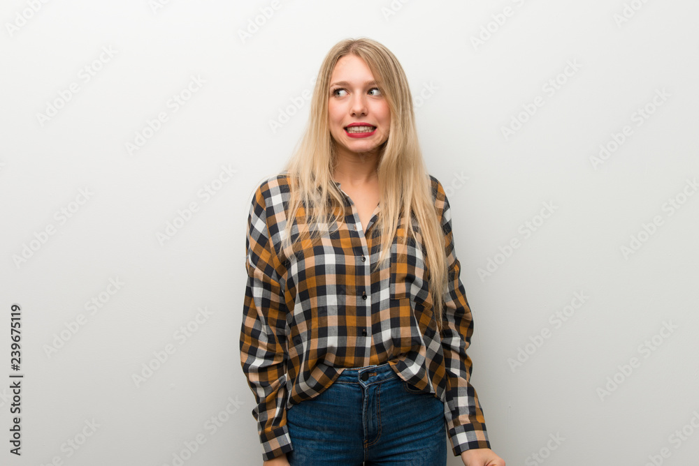 Blonde young girl over white wall is a little bit nervous and scared pressing the teeth Blonde young girl over white wall is a little bit nervous and scared pressing the teeth