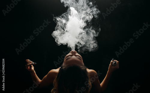 Redhead woman vaping electronic cigarette with smoke on black background closeup. Young woman smoking e-cigarette to quit tobacco. Vapor and alternative nicotine free smoking concept, copy space 