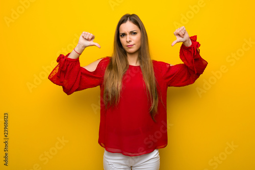 Young girl with red dress over yellow wall showing thumb down with both hands