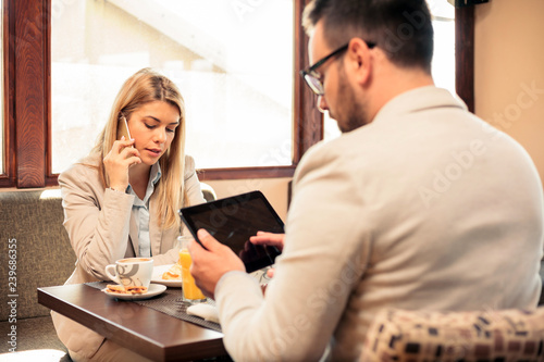 Two young male and female business partners having a meeting over breakfast in a cafe. Woman is talking on the phone