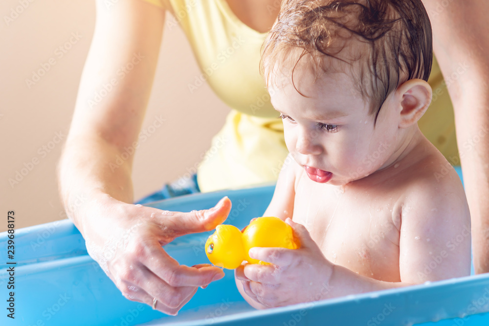 Mom washing little boy in a blue bath. Child is playing with a yellow duck. Hygiene and water treatment with the baby Mom washing little boy in a blue bath. Child is playing with a yellow duck. Hygiene and water treatment with the baby