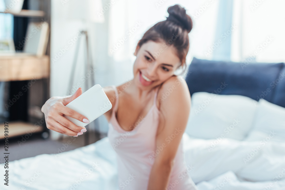 happy girl taking selfie on smartphone while sitting on bed during morning time at home happy girl taking selfie on smartphone while sitting on bed during morning time at home
