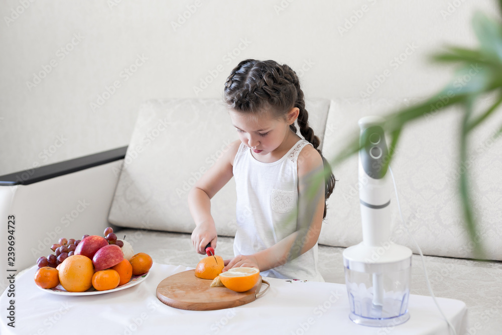 The child slices an orange with a knife. The girl cuts with a knife the fruit for making a cocktail.