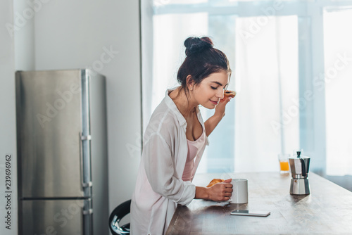 side view of girl having breakfast and checking smartphone near wooden table in kitchen