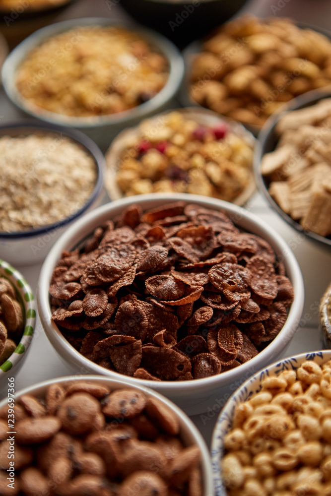 Close up and selective focus. Composition of different kinds cereals placed in ceramic bowls with cornflakes, granola, cereals and oatmeal. Flat lay, top view on white wooden table. Close up and selective focus. Composition of different kinds cereals placed in ceramic bowls with cornflakes, granola, cereals and oatmeal. Flat lay, top view on white wooden table.