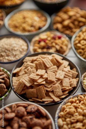 Close up and selective focus. Composition of different kinds cereals placed in ceramic bowls with cornflakes, granola, cereals and oatmeal. Flat lay, top view on white wooden table.