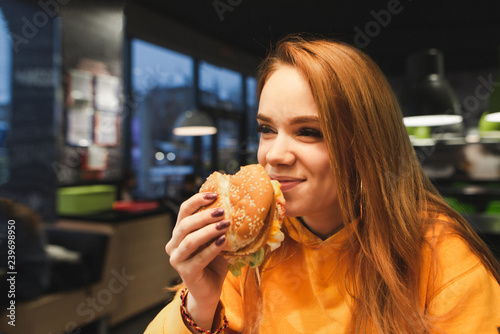 Close-up portrait of a girl holding a great tasty burger in her hand and laughing. A positive teen girl eats fast food at the evening in a cozy restaurant.