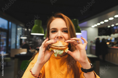Sweet girl holds a large tasty burger with two hands  bites and looks at the camera in close-up. Close-up portrait of a girl eats a burger  fast food at the restaurant background.