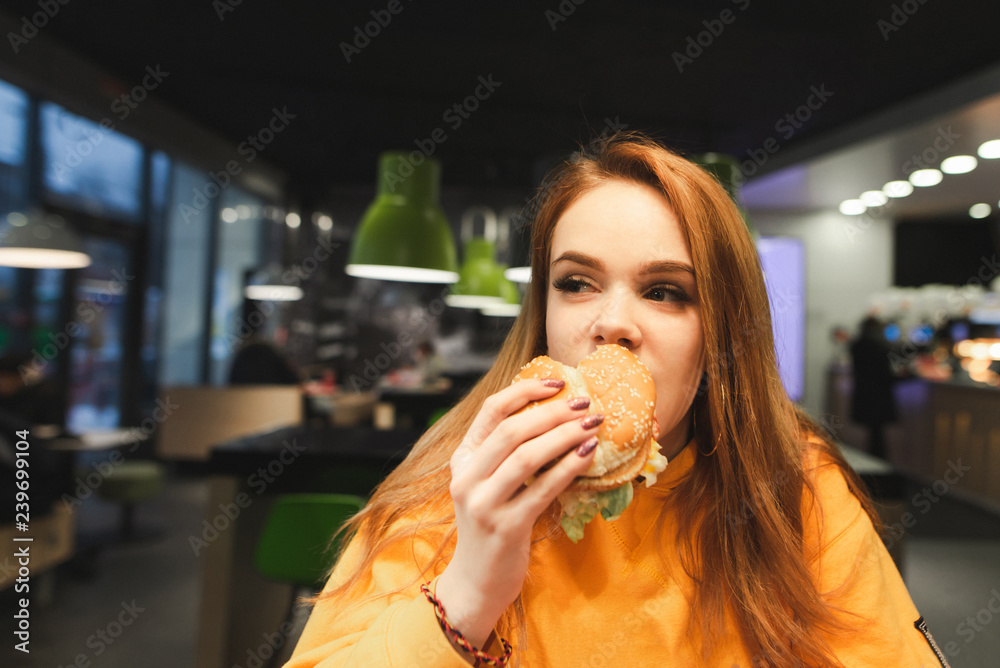 Sweet girl holds a large tasty burger with two hands, bites and looking sideways in close-up. Close-up portrait of a girl eats a burger, fast food at the restaurant background. Copyspace