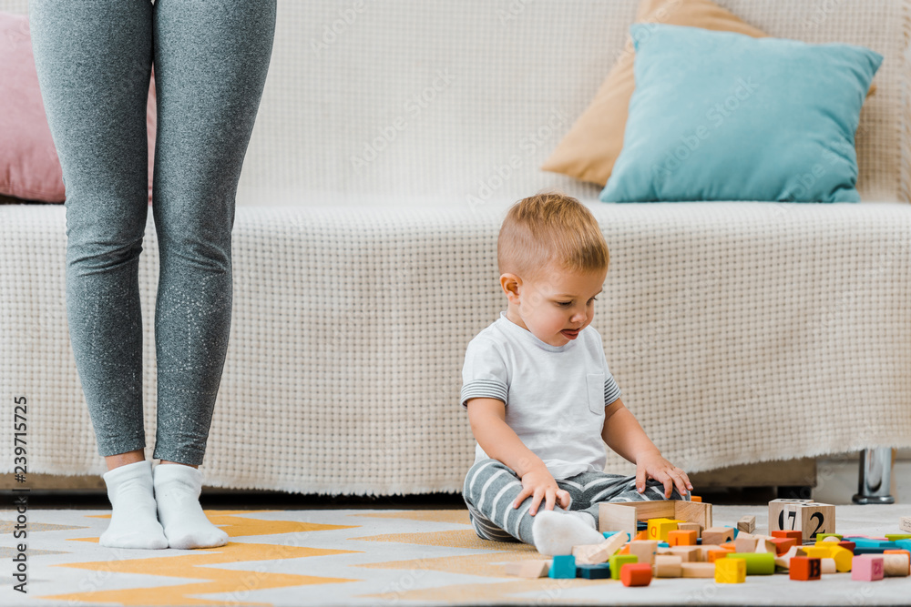Naklejka premium cute toddler boy playing with multicolored cubes and sitting on carpet at mom legs