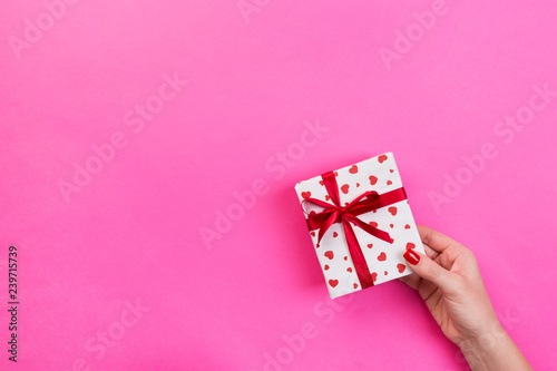Woman hands give wrapped valentine or other holiday handmade present in paper with red ribbon. Present box, red heart decoration of gift on pink table, top view with copy space for you design
