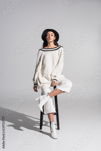 attractive african american girl in stylish white clothes and hat sitting on chair and looking at camera on white