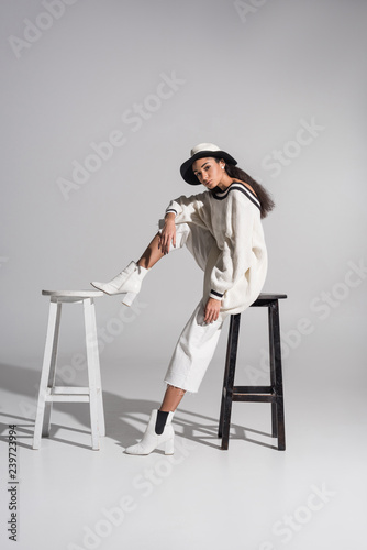 attractive african american girl in fashionable clothes and hat sitting on black and white chairs on white