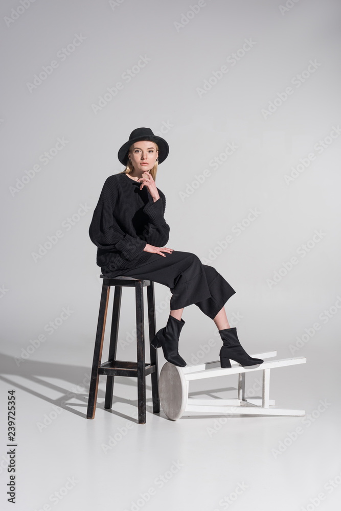 beautiful caucasian blonde woman in black clothes and hat sitting on chair and looking at camera on white beautiful caucasian blonde woman in black clothes and hat sitting on chair and looking at camera on white