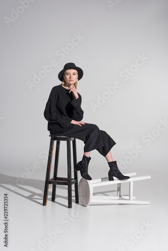 beautiful caucasian blonde woman in black clothes and hat sitting on chair and looking at camera on white