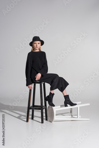 attractive caucasian blonde woman in black clothes and hat sitting on chair and looking at camera on white