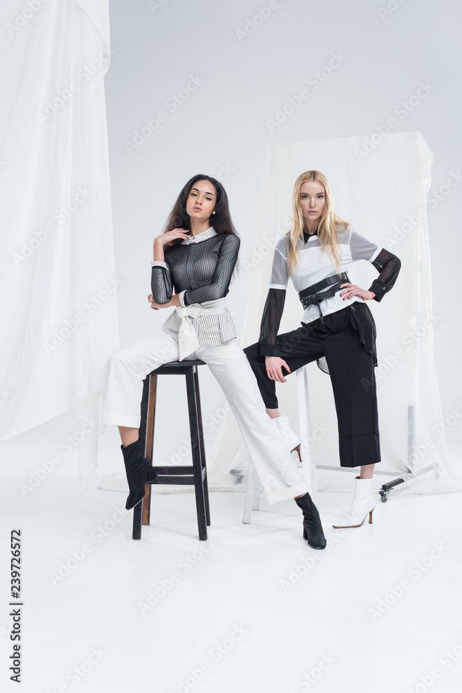 Fototapeta premium beautiful multiethnic women in fashionable black and white clothes posing on chairs on white