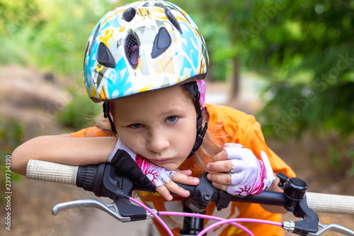 Cute little sad girl in a helmet with a bike in the summer