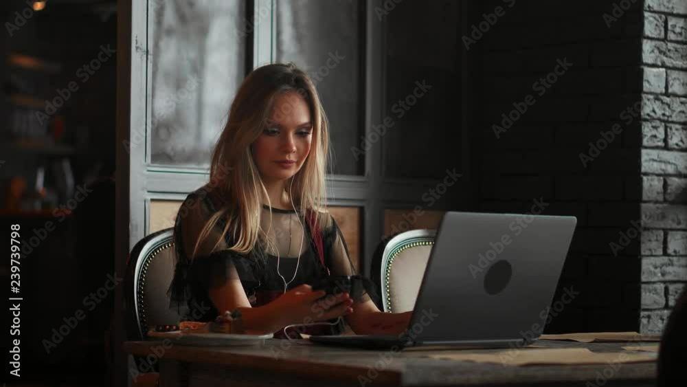 Young woman sitting in coffee shop at wooden table, drinking coffee and using smartphone.On table is laptop. Girl browsing internet, chatting, blogging. Female holding phone and looking on his screen. Young woman sitting in coffee shop at wooden table, drinking coffee and using smartphone.On table is laptop. Girl browsing internet, chatting, blogging. Female holding phone and looking on his screen.