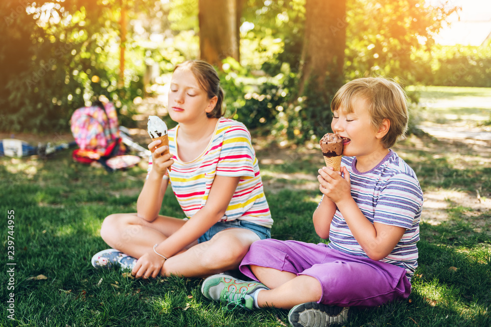 Two funny kids eating ice cream in summer park, sitting on the grass, wearing colorful stripe t-shirt Two funny kids eating ice cream in summer park, sitting on the grass, wearing colorful stripe t-shirt