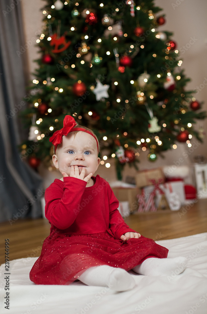 Little girl with blue eyes in a red dress sitting near the Christmas tree. Happy baby. Christmas homeliness. Holidays. Winter. Little girl with blue eyes in a red dress sitting near the Christmas tree. Happy baby. Christmas homeliness. Holidays. Winter.