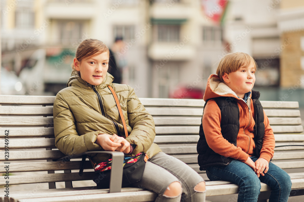 Outdoor portrait of two kids wearing warm jackets, sitting on the bench, spring fashion for children
