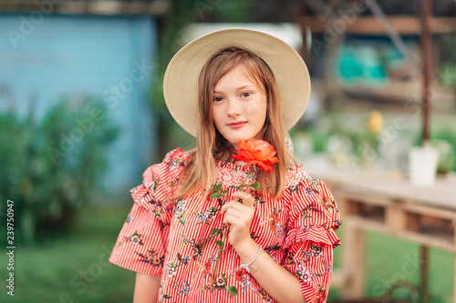 Summer portrait of pretty little girl wearing red stripe dress, big straw hat, holdin rose flower