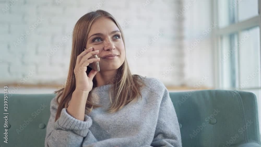 Attractive young woman with blond hair wearing gray is sitting on her couch at home, smiling, drinking tea and talking on smartphone Handheld slow motion medium shot Attractive young woman with blond hair wearing gray is sitting on her couch at home, smiling, drinking tea and talking on smartphone Handheld slow motion medium shot