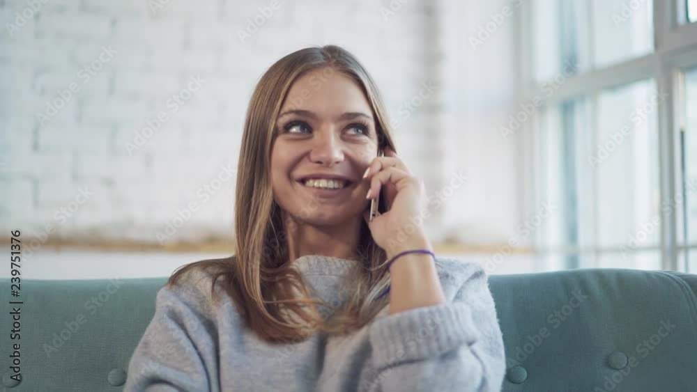 Smiling young blonde woman wearing gray sweater sitting on green sofa at home and talking on smartphone Slider slow motion medium shot Smiling young blonde woman wearing gray sweater sitting on green sofa at home and talking on smartphone Slider slow motion medium shot