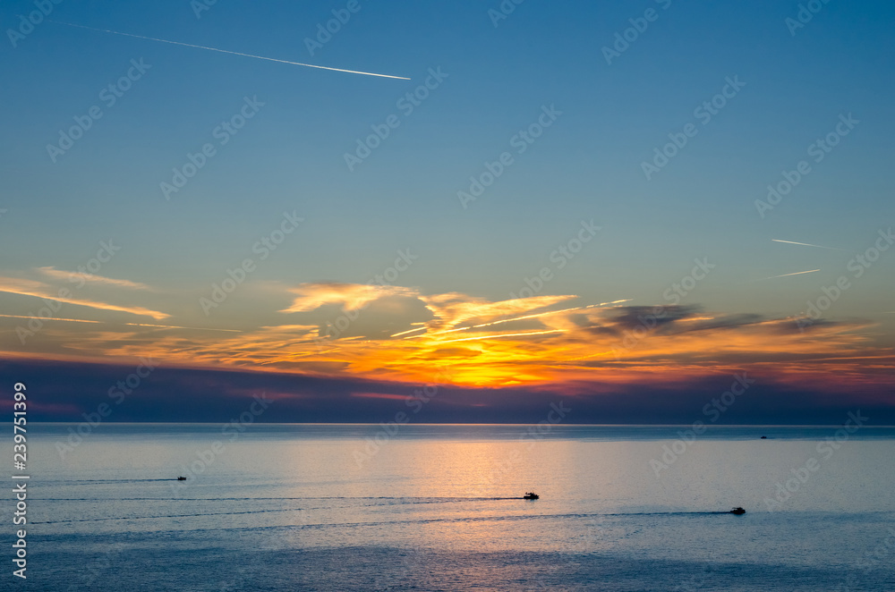 Sunset under the sea with moving pleasure boats and vapour trails on twilight sky Sunset under the sea with moving pleasure boats and vapour trails on twilight sky