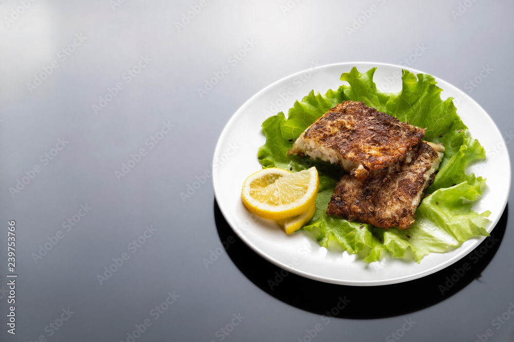 Two pieces of grilled fish fillet with the fresh leafs of lettuce and lemon on white plate on black background. Top view.